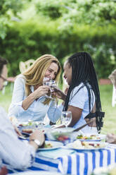 Glückliche Freundinnen stoßen mit Weingläsern am Esstisch während einer Gartenparty an - MASF14962
