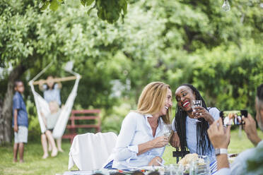 Man with mobile phone photographing smiling women having wine in garden party at weekend - MASF14965