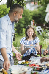 Girl holding food plate while sitting at table with family in backyard - MASF14974