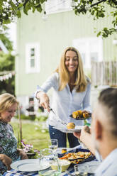 Frau serviert gegrillte Maiskolben an Freunde am Esstisch bei einer Party im Hinterhof - MASF14977