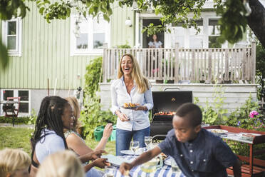 Glückliche reife Frau steht mit gegrilltem Essen Teller, während Freunde sitzen am Tisch im Hof während des Wochenendes - MASF14978