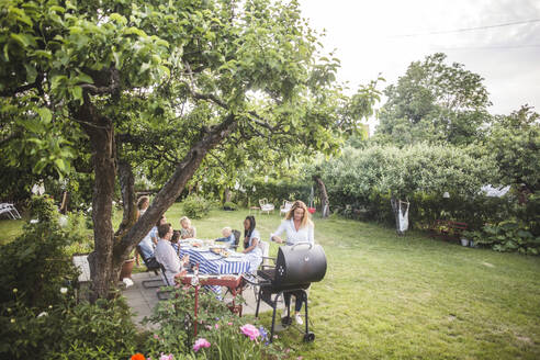 Frau bereitet Essen auf dem Grill zu, während die Familie am Tisch im Hinterhof sitzt - MASF14983