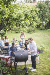 Hoher Blickwinkel des Mannes, der einen Grill mit einem Jungen vorbereitet, während Familie und Freunde am Tisch im Hinterhof sitzen - MASF14994