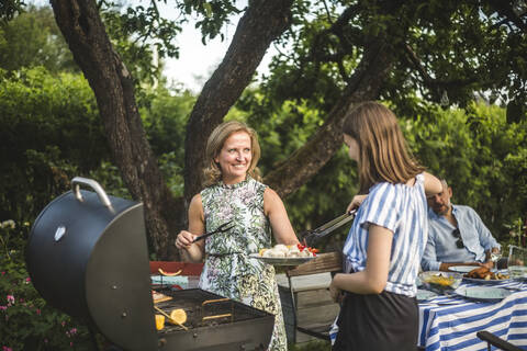 Lächelnde Mutter sieht ihre Tochter an, die bei der Zubereitung von Essen am Grill im Garten während einer Wochenendparty hilft, lizenzfreies Stockfoto