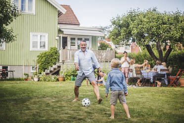 Vater spielt mit seinem Sohn Fußball, während Freunde sich am Tisch im Hinterhof amüsieren - MASF15006