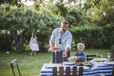 Mature father directing son with food plate while pointing at table in yard - MASF15014