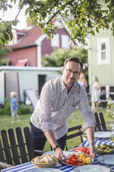Portrait of mature man placing food plate on table in backyard while preparing for garden party - MASF15016