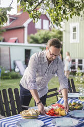 Man arranging food plate on table for garden party in backyard during summer weekend - MASF15017