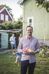 Porträt eines lächelnden reifen Mannes, der einen Teller mit Essen hält, während er im Hinterhof steht - MASF15019