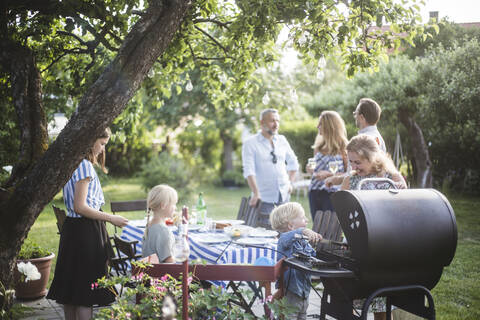 Junge, der mit seiner Mutter Essen auf dem Grill zubereitet, während die Familie am Esstisch im Hinterhof eine Party feiert, lizenzfreies Stockfoto