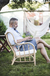Seitenansicht eines reifen Mannes, der auf einem Stuhl sitzend im Hinterhof an einem Laptop arbeitet - MASF15033