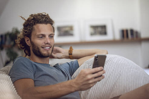 Lächelnder junger Mann, der sich zu Hause im Sitzsack entspannt und sein Handy benutzt - MCF00540