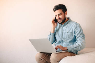 Glücklicher Mann, der mit einem Smartphone spricht, während er zu Hause einen Laptop benutzt - CAVF69464