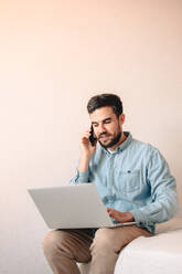 Man talking on smart phone while using laptop computer sitting at home - CAVF69465