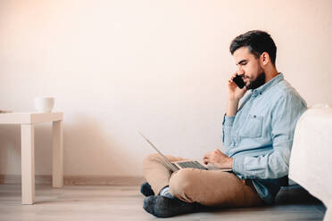 Man talking on smart phone using laptop sitting on floor against wall - CAVF69466