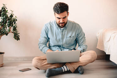 Junger Mann benutzt einen Laptop, während er zu Hause auf dem Boden sitzt - CAVF69468