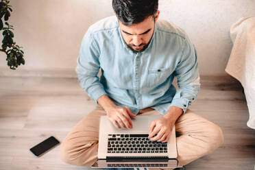 Junger Mann benutzt einen Laptop, während er zu Hause auf dem Boden sitzt - CAVF69469