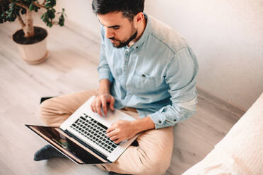 Ernster Mann, der einen Laptop benutzt, während er zu Hause auf dem Boden sitzt - CAVF69470