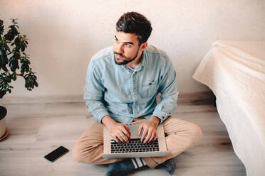 Mann benutzt einen Laptop, während er zu Hause auf dem Boden sitzt - CAVF69471