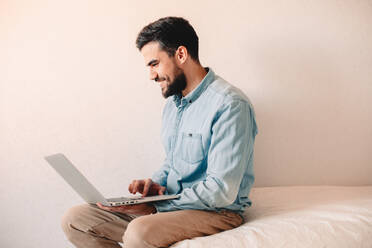 Glücklicher Mann, der einen Laptop benutzt, während er zu Hause an der Wand sitzt - CAVF69476