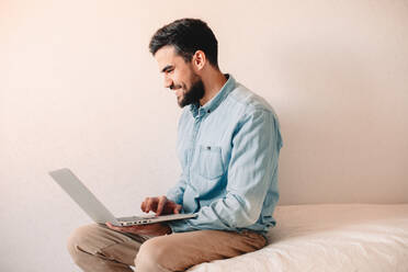 Happy man using laptop computer while sitting at home against wall - CAVF69476