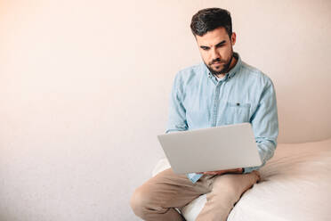 Junger Mann benutzt einen Laptop, während er zu Hause an der Wand sitzt - CAVF69478