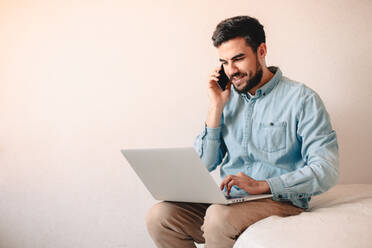 Happy businessman talking on smart phone while using laptop at home - CAVF69479