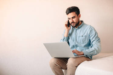 Mann spricht mit einem Smartphone, während er zu Hause einen Laptop benutzt - CAVF69480