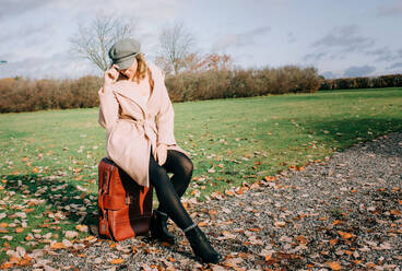 Woman sitting on a suitcase waiting on a country road in fall - CAVF69555