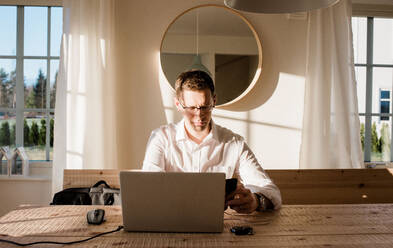 Mann arbeitet von zu Hause aus und schaut konzentriert auf sein Telefon und seinen Laptop - CAVF69573