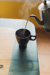 Tee aus dem Kessel in eine Tasse auf einem Holztisch gießend - CAVF69609