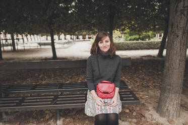 Porträt einer lächelnden Frau mit Handtasche auf einer Bank im Park - CAVF69642