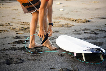 Niedriger Ausschnitt eines Mannes, der auf nassem Sand am Strand stehend sein Surfbrett festbindet - CAVF69653