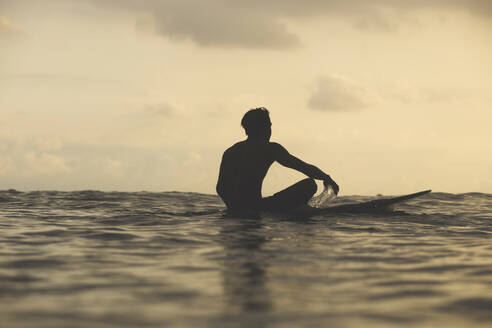 Rückansicht eines Mannes, der auf einem Surfbrett im Meer sitzt, gegen einen bewölkten Himmel bei Sonnenuntergang - CAVF69690