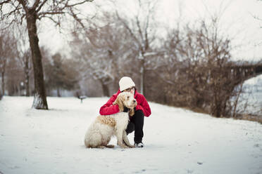 Frau mit Hund, die wegschaut, während sie auf einem schneebedeckten Feld hockt - CAVF69746