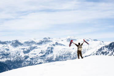 Mann mit Rucksack, stehend auf Berggipfel, winkend mit seinen Schneeschuhen, Rückansicht, Bundesland Salzburg, Österreich - HHF05591