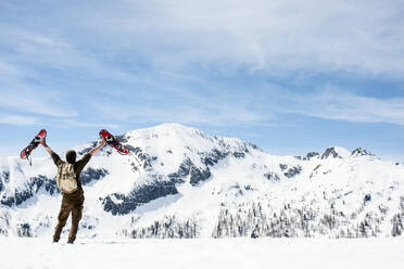 Mann mit Rucksack, stehend auf Berggipfel, winkend mit seinen Schneeschuhen, Rückansicht, Bundesland Salzburg, Österreich - HHF05592