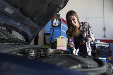 Junge Frau in Hemd und Handschuhen bei der Arbeit mit einem Auto in einer Reparaturwerkstatt, die den Motorölstand überprüft - ABZF02853