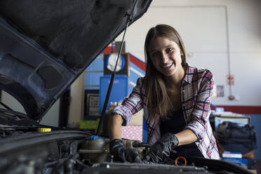 Junge lässige Frau, die in die Kamera lächelt, während sie einen Automotor repariert und im Autoservice arbeitet - ABZF02860