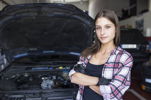 Lässige Frau in kariertem Hemd und Handschuhen, die die Arme verschränkt hält und in die Kamera lächelt, arbeitet im Autoservice, lizenzfreies Stockfoto