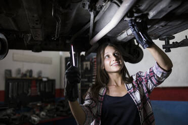 Lächelnde Frau mit Lampe, die ein Auto repariert, das unter dem Boden in einer Autoreparaturwerkstatt steht - ABZF02865