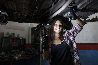 Casual woman with lamp standing under car bottom on leveling system and smiling at camera working in service - ABZF02866