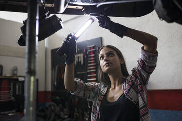 Brünette Frau im Hemd, die eine brennende Lampe hält und mit einem Auto arbeitet, das unter dem Boden in einer Autowerkstatt steht - ABZF02867