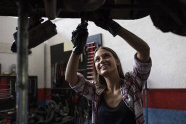 Junge Frau mit Handschuhen, die einen Schraubenschlüssel hält und ein Auto repariert, das unter dem Boden in einer Reparaturwerkstatt steht - ABZF02868