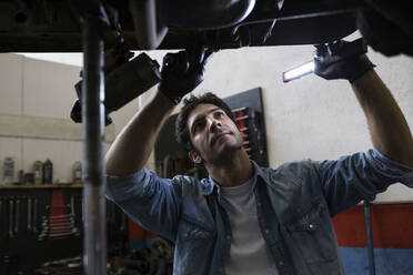 Mann im Hemd, der unter einem Auto steht und mit einem Werkzeug in einer Werkstatt arbeitet - ABZF02869