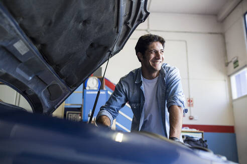 Lächelnder Mann, der mit dem Motor eines Autos in der Reparaturwerkstatt arbeitet und wegschaut - ABZF02876