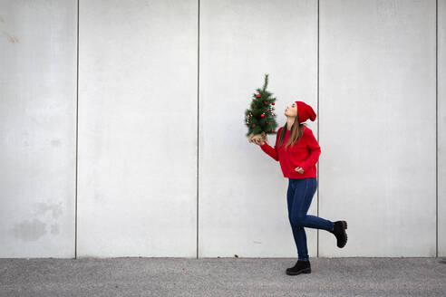 Frau mit rotem Pullover und lustiger Mütze, die einen künstlichen Weihnachtsbaum vor einer Wand hält - HMEF00685