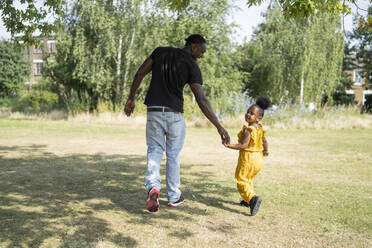 Rückansicht von Vater und Tochter, die Hand in Hand in einem Park spazieren gehen - FBAF00988