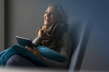 Lachende junge Frau, die in einem Sessel sitzt und ein Tablet benutzt - MOEF02648