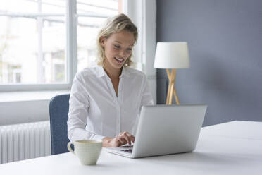 Lächelnde junge Geschäftsfrau mit Laptop am Schreibtisch im Büro - MOEF02684